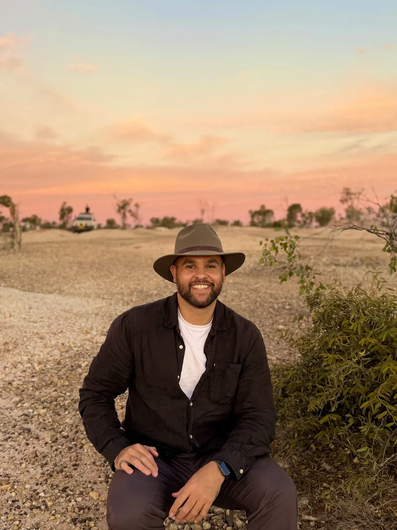 James Saunders seated outdoors in a wide-brim hat, with the outback at sunset behind him.