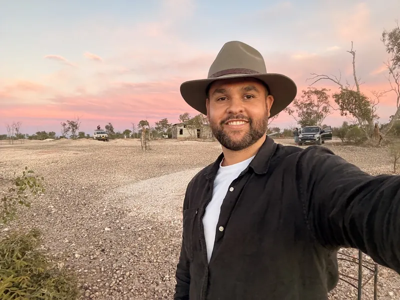 James Saunders in a wide-brim hat, smiling, with the outback at sunset behind him.