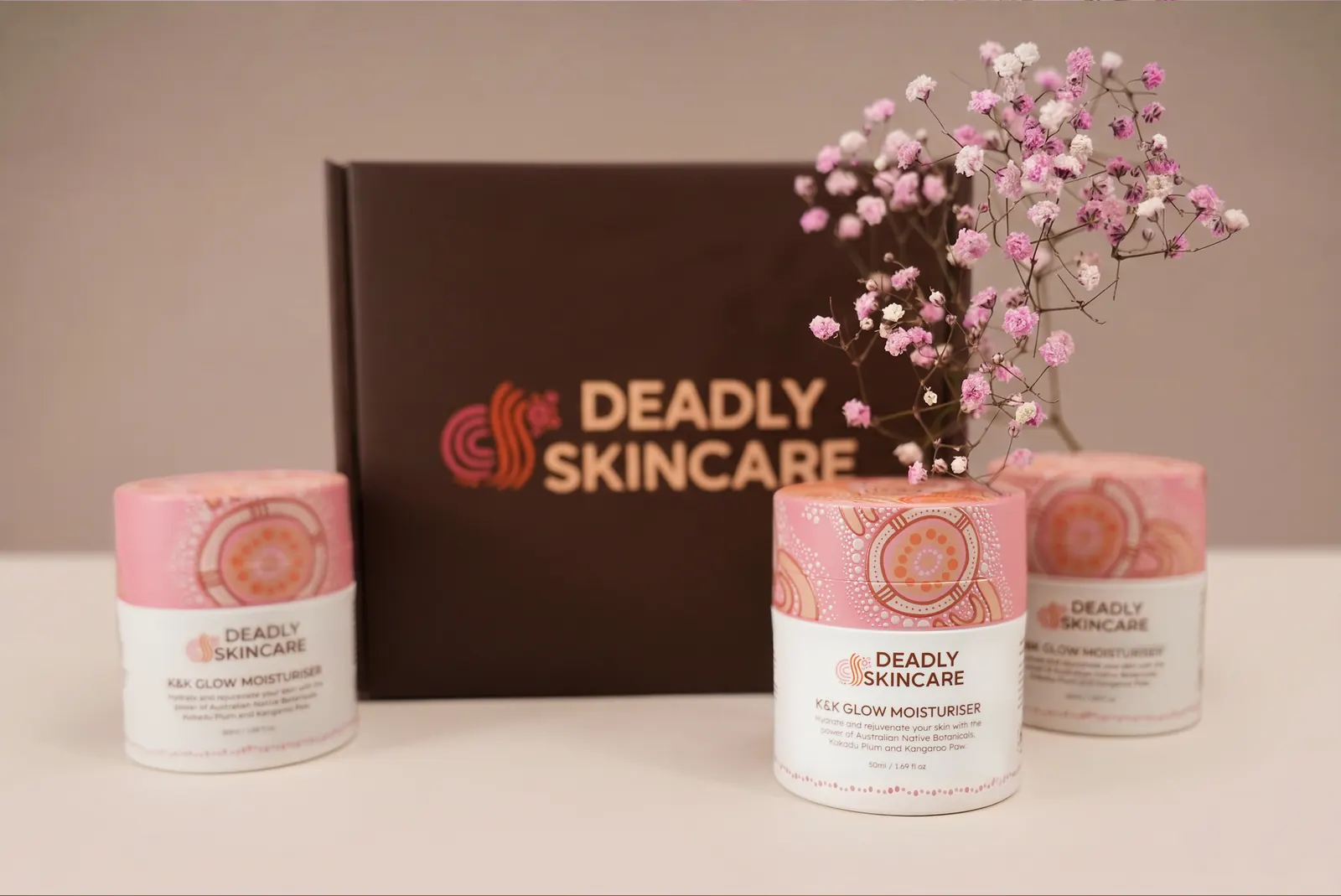 Deadly Skincare moisturiser jars in front of the brand gift box, with pink flowers.