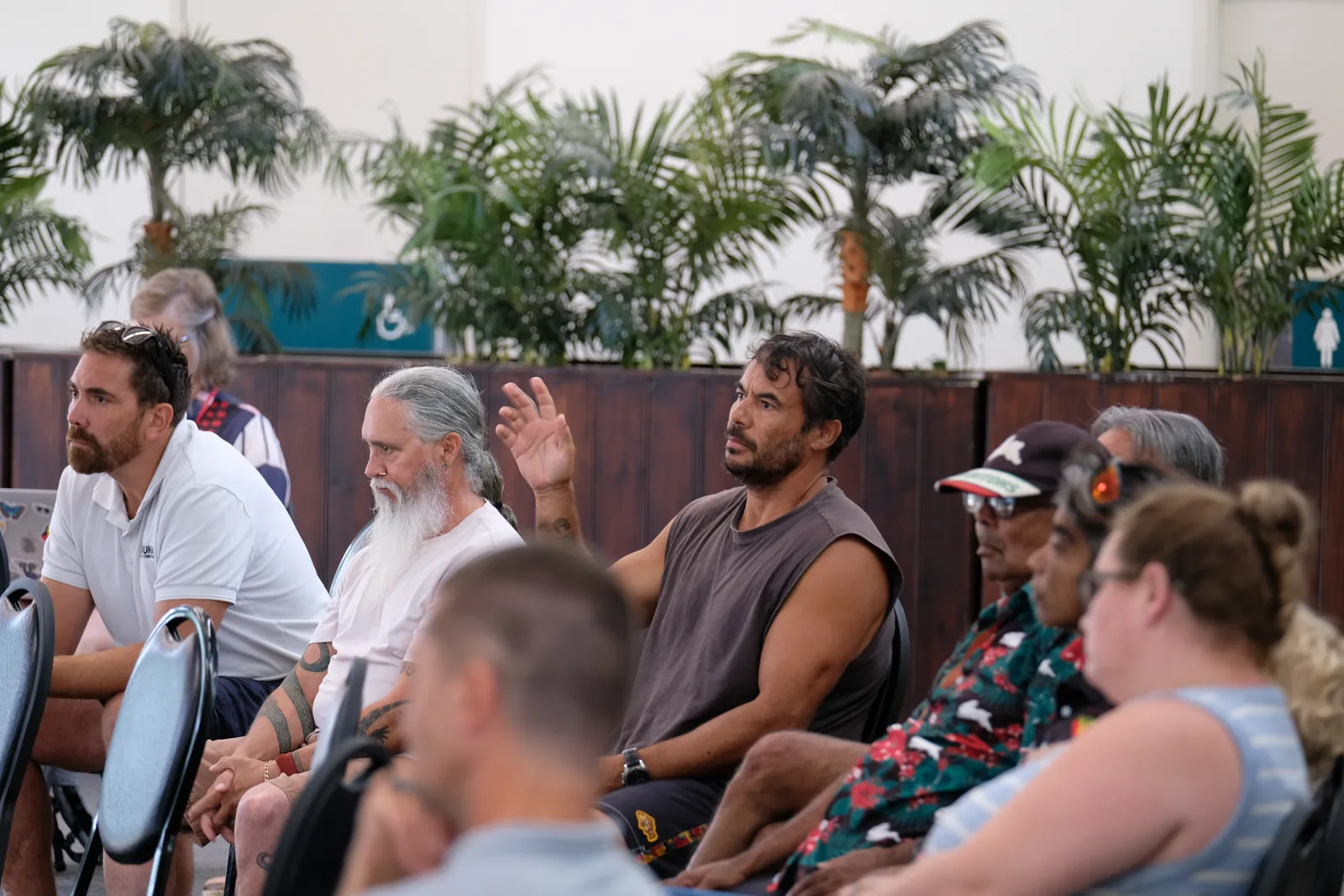 Community members seated together in a hall, listening as one man raises his hand to speak.