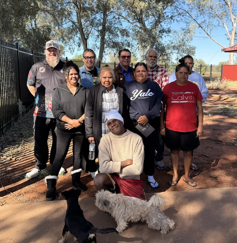 Community members outdoors on red earth, with two dogs at their feet.